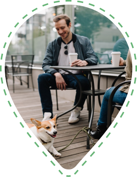 An image of two friends drinking coffee outside at a cafe and smiling at a Corgi sitting at their feet.